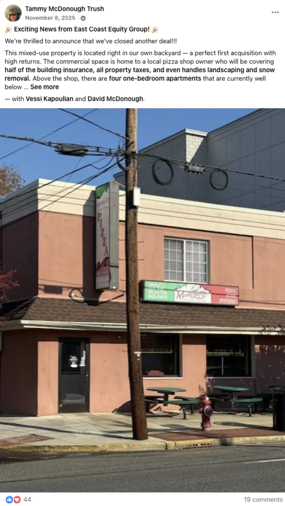 Front view of a small pizza shop with outdoor seating on a sidewalk, pinkish building and a utility pole in the foreground.